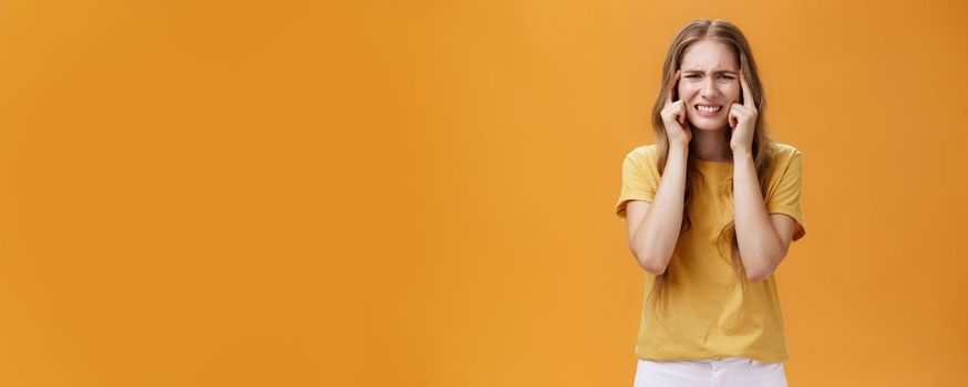 Girl gonna blow up from pressure feeling intense and unfocused squinting and frowning holding fingers on temples, clenching teeth having trouble to remember or feeling headache over orange wall.