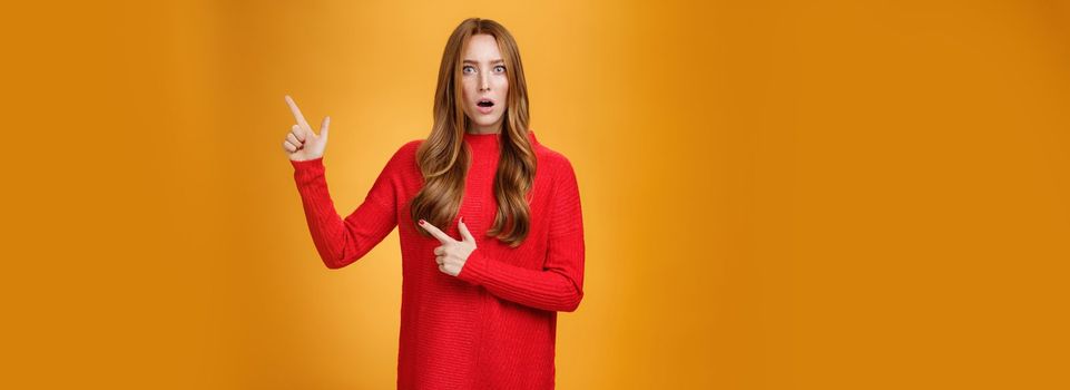 Shocked distressed and confused ginger girl with freckles and blue eyes in red sweater pointing at upper right corner with shook and surprise open mouth, raise eyebrows from stupor over orange wall.