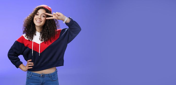 Lifestyle. Attractive feminine woman with curly hairstyle in hat and sweatshirt showing peace or victory sign around eye and smiling carefree having fun playing in yard with snow over blue background.