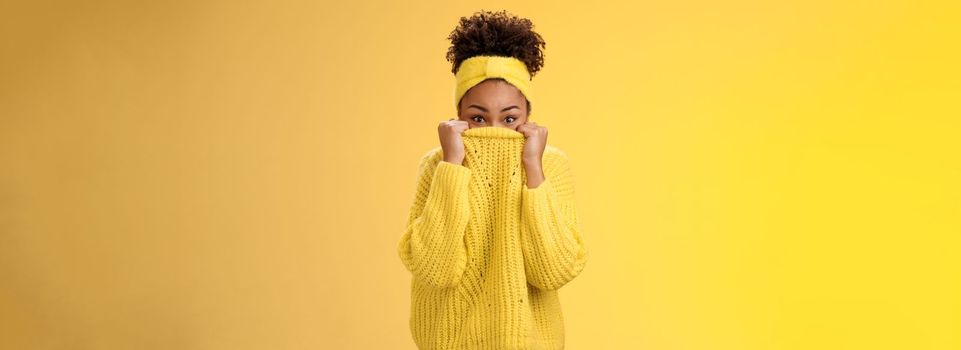 Charming cute silly young african-american modern girl. pulling sweater on face hiding peeking camera playfully having fun smiling hide-n-seek fool around mimicking standing yellow background.