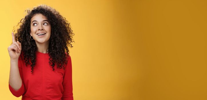 I got idea. Excited happy attractive woman with curly hair in red dress raising index finger in eureka gesture smiling amused, looking at upper left corner, sharing plan or suggestion over yellow wall.