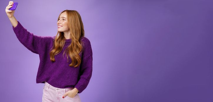 Attractive confident and stylish happy 20s woman with ginger hair standing carefree with joyful smile over purple background extending hand with smartphone as taking selfie happily. Technology and lifestyle concept