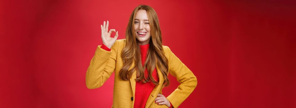 Okay got it. Portrait of carefree happy and confident good-looking charismatic ginger woman with freckles winking and showing tongue joyfully showing ok gesture, liking excellent work over red wall.