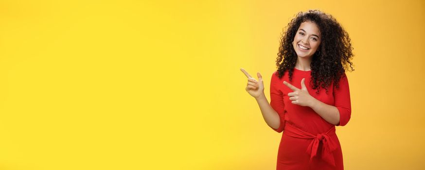 Friendly cute assertive female assistant in elegant red dress with curly hair tilting head flirty and smiling broadly, satisfied as pointing at upper left corner, recommending product over yellow wall.