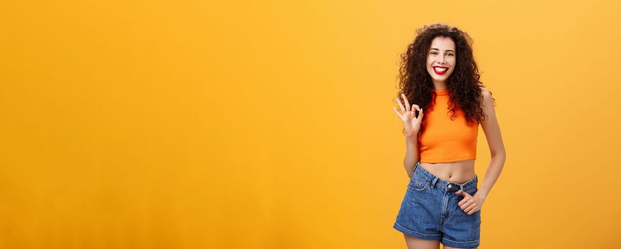 Charming young charistmatic woman. with curly hairstyle in cropped top and shorts showing okay or approval gesture smiling broadly liking awesome idea of friend where hang out over orange wall.