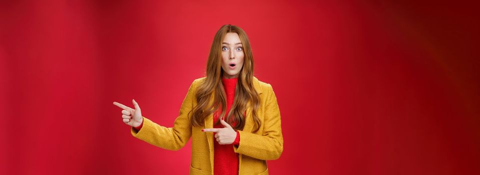 Speechless questioned and confused surprised redhead girl in yellow autumn coat pointing right open mouth curiously and staring at camera puzzled and clueless waiting explanation over red wall. Advertisement and fashion concept