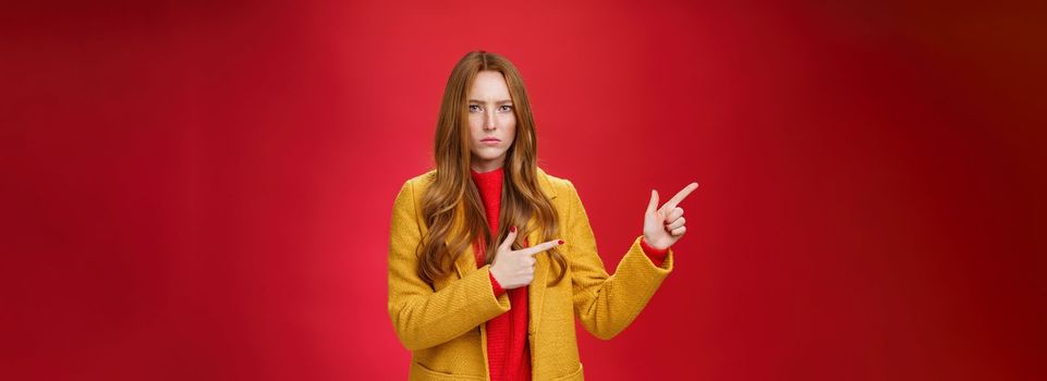Displeased offended and gloomy girlfriend asking question, pouting and frowning from insult pointing at upper left corner disappointed and upset posing dissatisfied against red background.