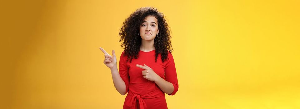 Waist-up shot of upset displeased girl with curly hair being down in dumps pointing at upper right corner with disappointed gloomy smile, feeling regret, sadness standing like loser over yellow wall.