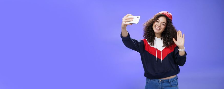 Studio shto of stylish cute female vlogger with curly hair in winter red hat smiling and waving hello at smartphone camera as recording video, making interesting content to post online over blue wall.