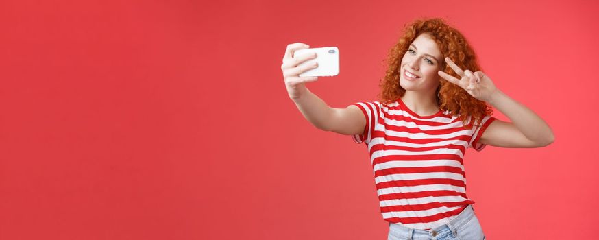 Stylish carefree good-looking redhead curly woman enjoy summer holidays show victory peace gesture eye tilt head cute record video hold smartphone taking selfie red background. Copy space