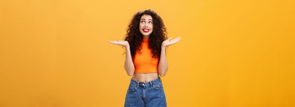 Silly and clueless carefree cute girl. with curly hairstyle in cropped top and shorts shrugging with palms spread aside smiling unaware and gazing at upper right corner uncertain and uninvolved.