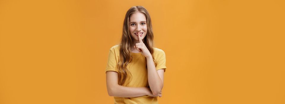 Girl wants keep secret betweet us. Portrait of charming good-looking slim pretty and young woman with long wavy hair and tattoo showing shh gesture with index finger over mouth against orange wall.