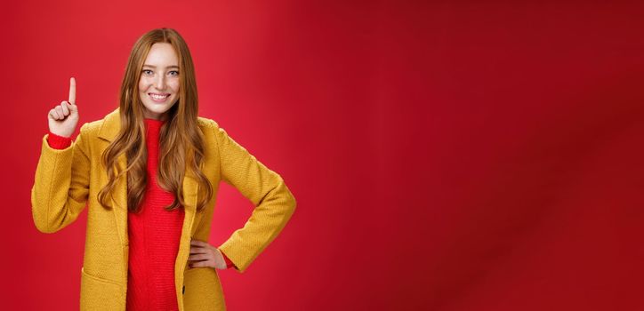 Girl telling first reason use her advice. Cute and friendly energized young redhead woman in yellow coat raising hand showing number one and smiling broadly at camera against red background.