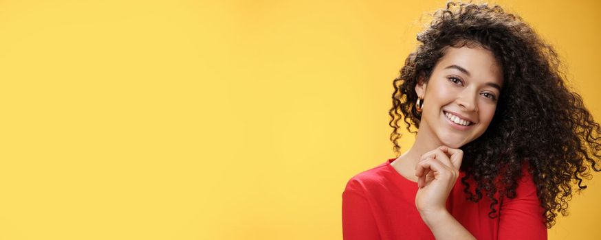 Lifestyle. Close-up shot of stylish and happy bright curly-haired female in red dress tilting head sensually touching chin with finger and smiling broadly making flirty gazed at camera over yellow background.
