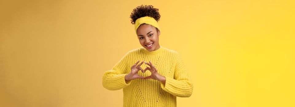 Lovely tender stylish african-american girl curly hairstyle headband sweater showing heart love gesture gladly smiling flirty tilting head express sympathy passion desire, posing yellow background.