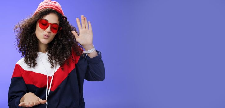 Lifestyle. Studio shot of cool and stylish dj girl in red beanie and sunglasses raising palm as enjoying cool music and dancing to rhythm folding lips having fun at awesome party posing chill over blue wall.