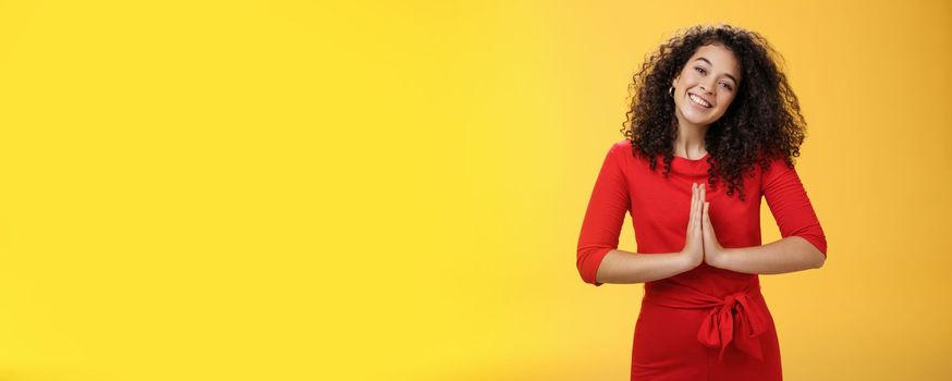 Welcome come inside. Portrait of friendly and polite good-looking female host in red dress with curly hair holding hands in namaste gesture tilting head and smiling as inviting guests in asian style.