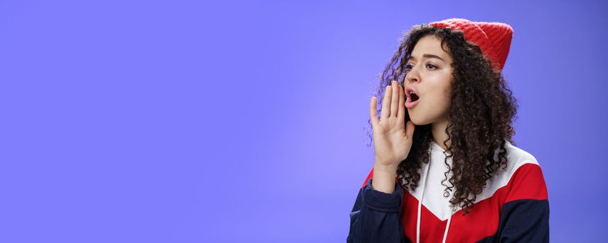 Portrait of worried sister calling sibling outdoors open mouth looking left seriously and holding palm near lips as shouting name searching someone, posing concerned over blue background.