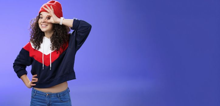 Okay girl agrees. Friendly-looking happy and charming young curly-haired girl in warm beanie showing ok gesture or circle over eye as peeking at camera and smiling broadly with joy over blue wall.