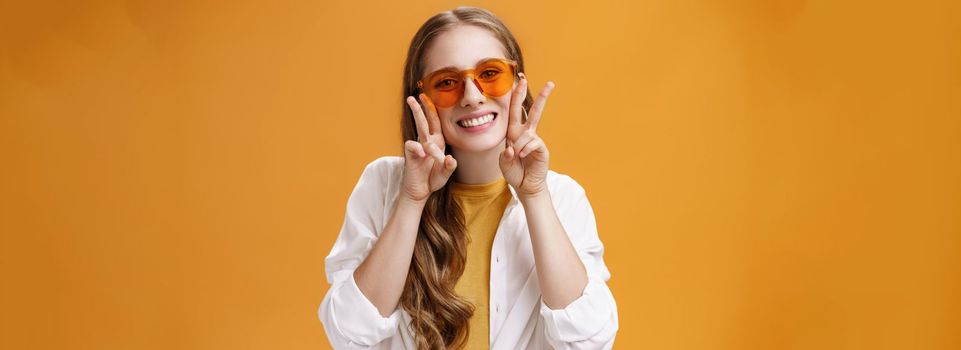 Stylish enthusiastic and charismatic young party girl in trendy sunglasses and white blouse over t-shirt showing peace gestures near face and smiling cute at camera against orange background. Copy space