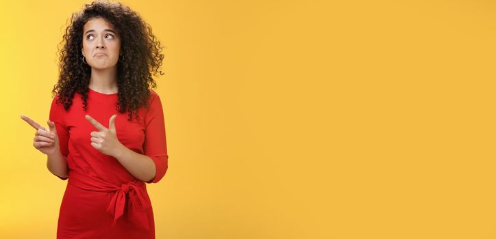 Gloomy upset and sad cute silly curly-haired girl in red dress pursing lips down in unhappy smile frowning from regret, missing chance or offer pointing at upper left corner over yellow background.
