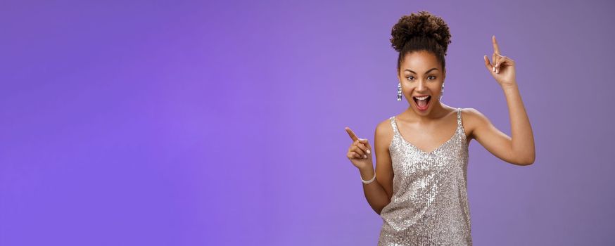 Sassy good-looking african-american woman in trendy glamour silver shiny dress index fingers raised up smiling cheeky joyfully celebrating great lucky news dancing grinning, standing blue background.