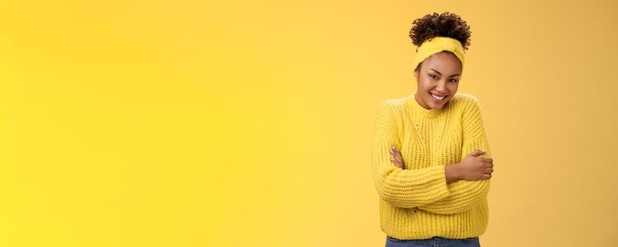 Tender delicate young african-american cheerful girlfriend curly hairstyle headband sweater hugging herself embracing gladly smiling camera feel soft comfortable, standing yellow background warm.