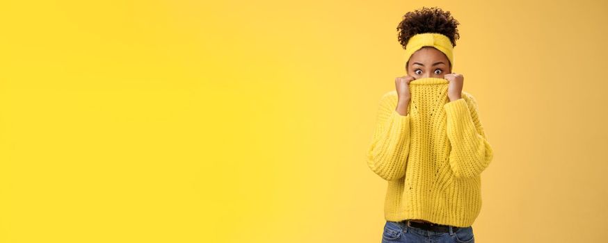 Shocked afraid stunned young beautiful african-american trendy girl curly afro hairstyle pull sweater nose look from collar widen eyes amazed surprised frightened unexpected loud noise, yellow wall.