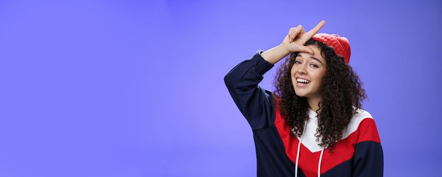 Girl triumphing and mocking friend as winning showing loser gesture on forehead and laughing having fun standing pleased and happy over blue background, smiling at camera joyfully.