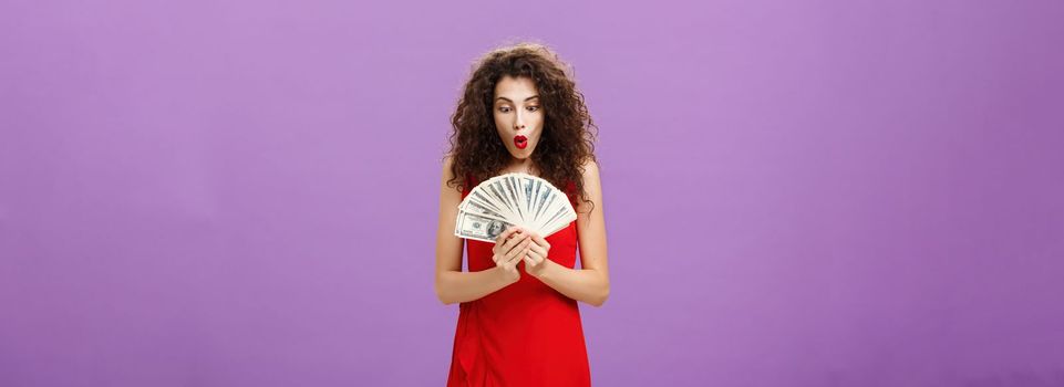 Portrait of surprised and amazed speechless woman folding lips making wow sound while holding and looking at lots of money being rich and successful receiving lots of cash over purple background. Finance concept