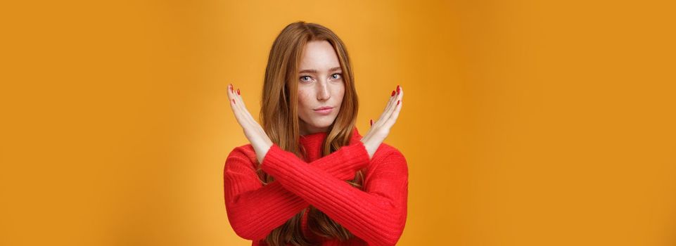 Indoor shot of self-assured attractive powerful woman with red hair and freckles cross arms in no and prohibition gesture forbidding make action standing confident over orange wall. Body language concept