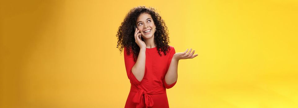 Carefree talkative cute woman with curly hair in red dress having char with friend on mobile phone holding smartphone near ear, gesturing as discussing exciting news, looking at upper left corner.