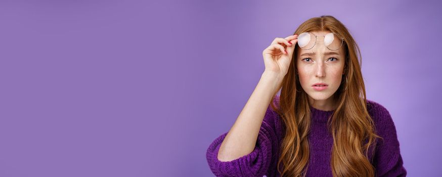 Waist-up shot of intense confused and unsure cute redhead female cannot look without glasses having bad sight taking off eyewear and squinting at camera uncertain, unable to read over purple wall.