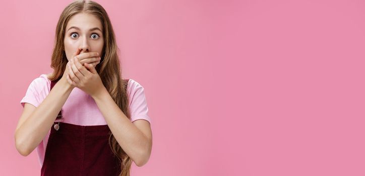 Lifestyle. Waist-up shot of shocked speechless pretty young girl in overalls with make-up covering mouth with palms from overreacting on terrible news witnessing accident nervously over pink background.