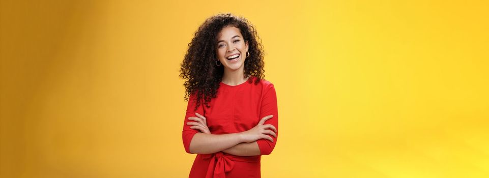 Self-assured happy enthusiastic curly-haired female reporter in cute red dress laughing carefree, having fun tilting head amused and holding hands crossed over body in confident pose over yellow wall.