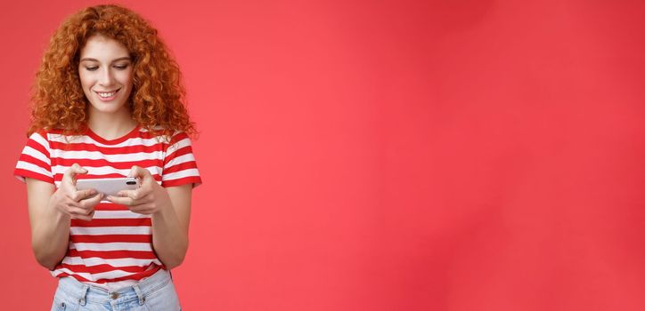 Lifestyle. Redhead girl fool around waiting queue dentist playing awesome smartphone game hold phone horizontal tap cellphone screen look telephone display smiling delighted entertained standing red background.