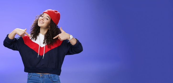 Lifestyle. Proud and self-satisfied happy young energized stylish female model in warm beanie brag about own achievements as pointing at herself raising head and smiling produly posing delighted over blue wall.