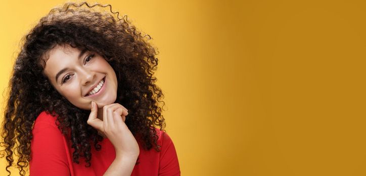 Close-up shot of charming flirty and silly curly-haired young woman making faces as trying get what wants, smiling tilting head on shoulder and touching lip standing cute over yellow background. Romance, people and beauty concept