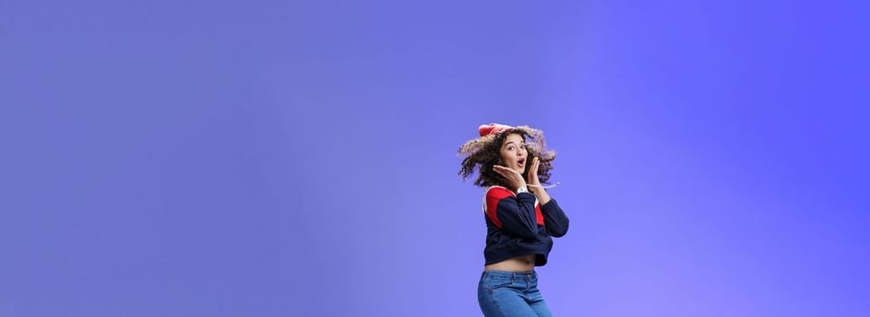 Lifestyle. Surprised and amazed amused good-looking woman with curly hair in winter beanie and sweatshirt open mouth from joy and amazement jumping over blue background having fun feeling playful.