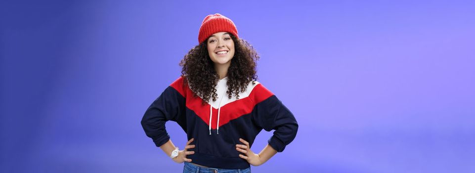 Lifestyle. Confident ad energized cute european woman with curly hairstyle in warm beanie and stylish sweatshirt holding hands on waist and smiling friendly at camera as ready to go out and have fun outside.