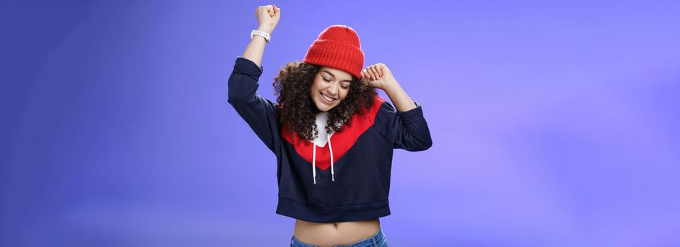 Girl celebrating favorite season in year. Portrait of carefree and joyful dancing woman with curly hair in cute red hat lifting hands in dance movements smiling enjoying music and holidays.