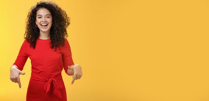 Waist-up shot of assertive charming and happy young woman with curly hairstyle laughing joyfully, smiling and pointing down as showing cool place to check out over yellow background. Advertisement concept