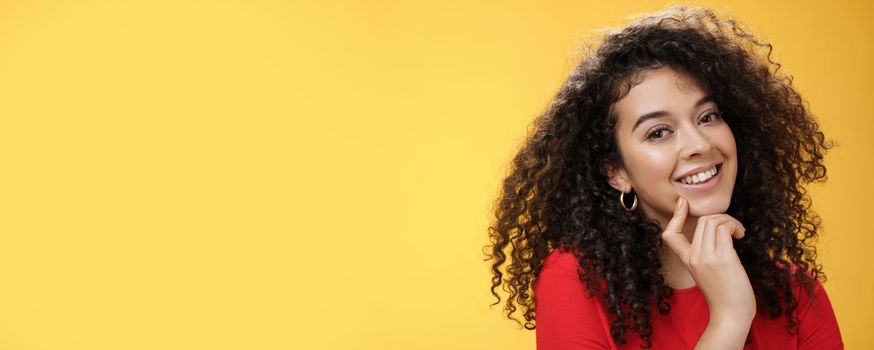 Lifestyle. Headshot of cute silly and tender feminine romantic woman with curly hairstyle touching lip with index finger making eyes at camera and smiling as using seduction skills over yellow background.