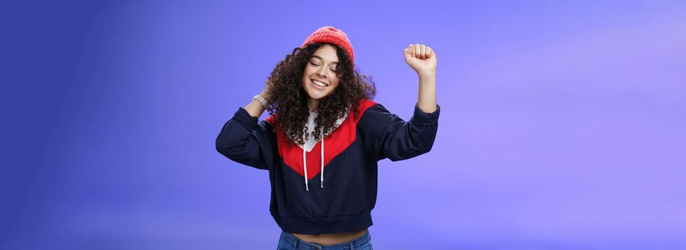 Girl delighted and carefree dancing over blue background with closed eyes and tender smile moving raised hands in rhythm of music, dreaming having positive mood wearing warm winter hat and sweatshirt.