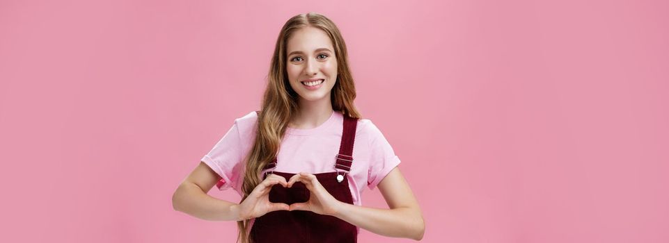 Girl loves family. Kind charming young woman in overalls with small tattoo on arm showing heart gesture over body and smiling lovely at camera expressing tender and cute attitude over pink wall.