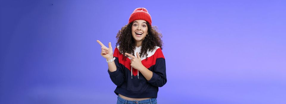 Lifestyle. Enthusiastic friendly-looking attractive young 20s woman with curly hair in warm beanie and sweatshirt smiling open mouth amazed and delighted as pointing at upper right corner over blue background.