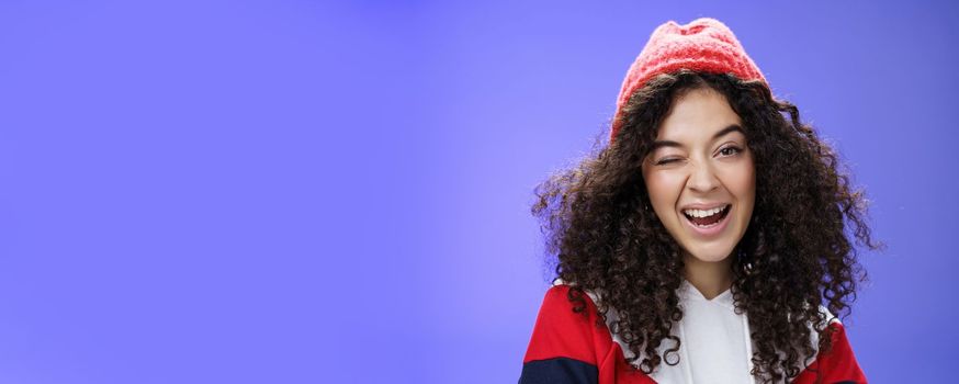 Charming cute woman hinting to go play snowballs with her winking joyfully as inviting us, smiling broadly wearing stylish red beanie and sweatshirt as posing against blue background.