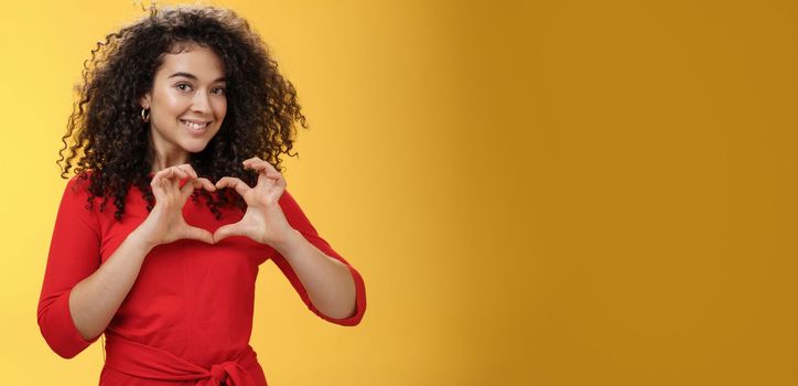 Lifestyle. Waist-up shot of romantic and cute pretty girlfriend with curly hair in red dress showing heart sign over chest and smiling broadly confessing in admiration and love posing over yellow background.