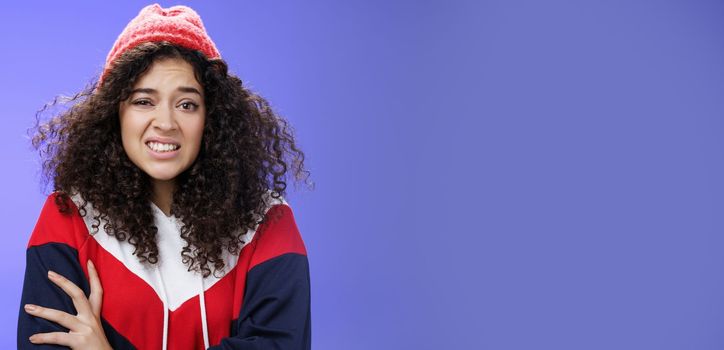Woman with curly hair in winter beanie feeling uncomfortable and discomfort clenching teeth and frowning intense as hugging herself insecure and awkward, unwilling to say cruel rejection, feel awkward.