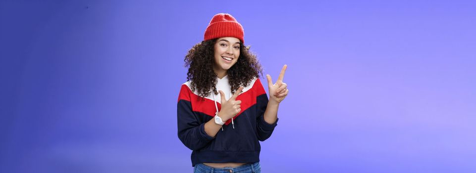 Lifestyle. Portrait of friendly good-looking woman with curly hair in red warm beanie pointing at upper left corner and smiling delighted as promoting cool copy space against blue background.